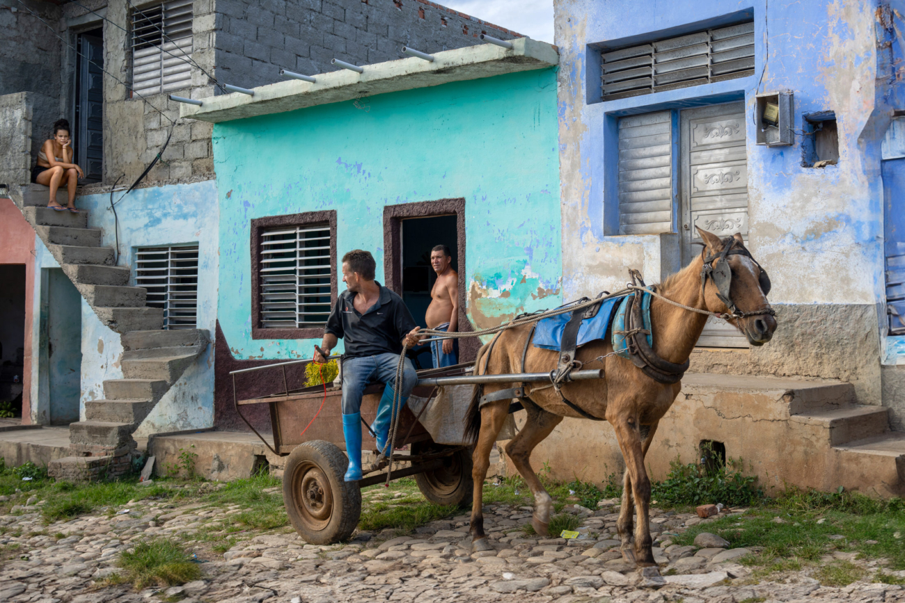 Cuba Street Photography Roadtrp | 14 Day Guide with Mark Seymour 9 “Candid street scene in rural Cuba with a man riding a horse-drawn cart, a woman sitting on outdoor steps, and colourful weathered buildings in the background.”