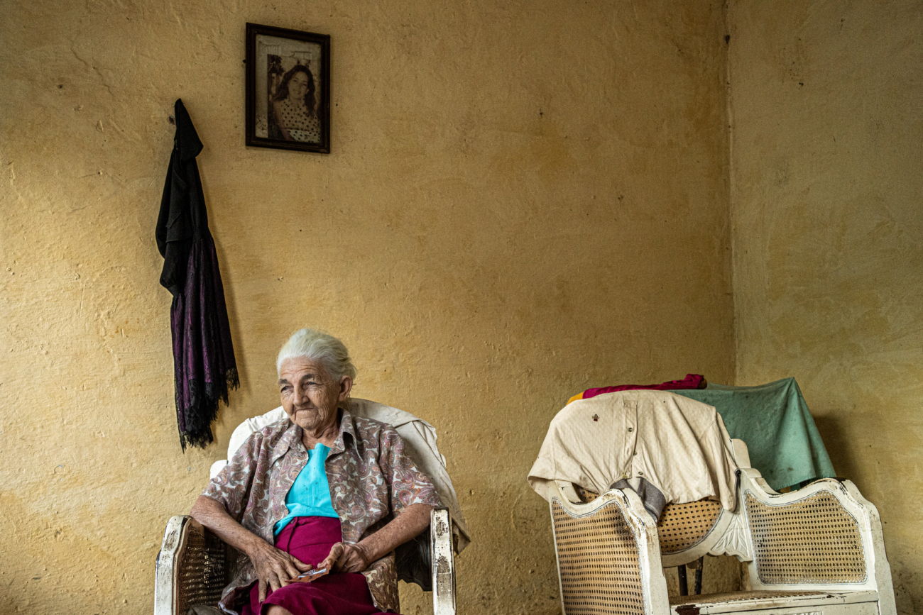 Cuba Street Photography Roadtrp | 14 Day Guide with Mark Seymour 10 “Elderly Cuban woman sitting calmly in a worn wicker chair inside a simple yellow room with family photos on the wall.”