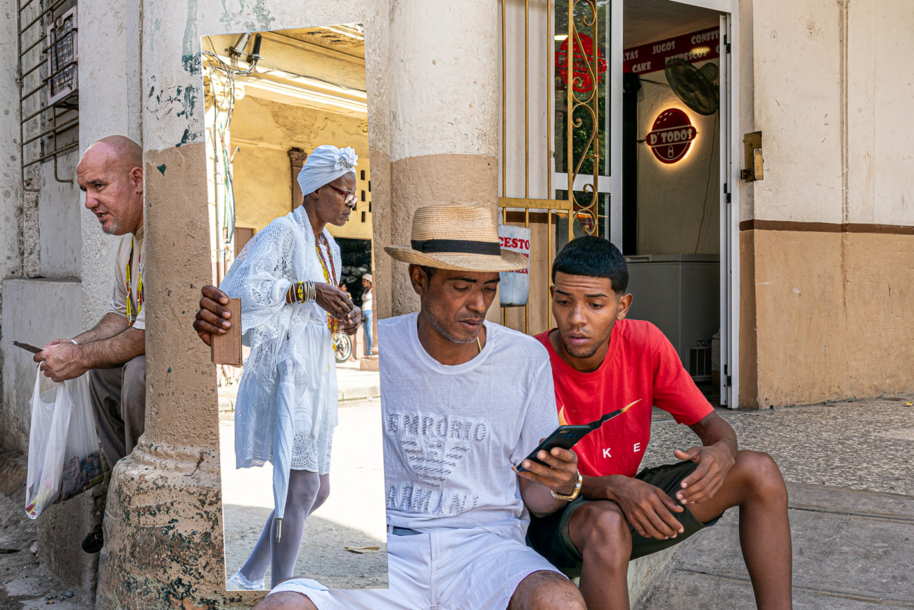 Cuba Street Photography Roadtrp | 14 Day Guide with Mark Seymour 2 “Street moment in Havana with two men checking a mobile phone as a woman in traditional white clothing passes behind them.”