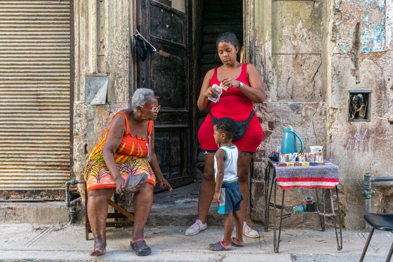 Cuba Street Photography Roadtrp | 14 Day Guide with Mark Seymour 14 “Street vendor in a red dress preparing items for sale as an elderly woman and a young boy stand beside her in a worn Havana doorway.”