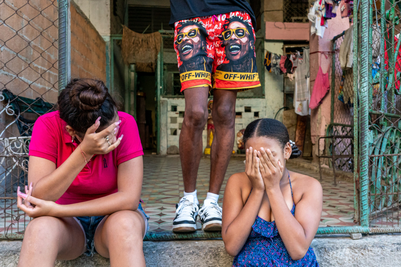 Cuba Street Photography Roadtrp | 14 Day Guide with Mark Seymour 4 “Two Cuban girls sitting on a doorstep covering their faces while a man in vibrant shorts stands behind them.”