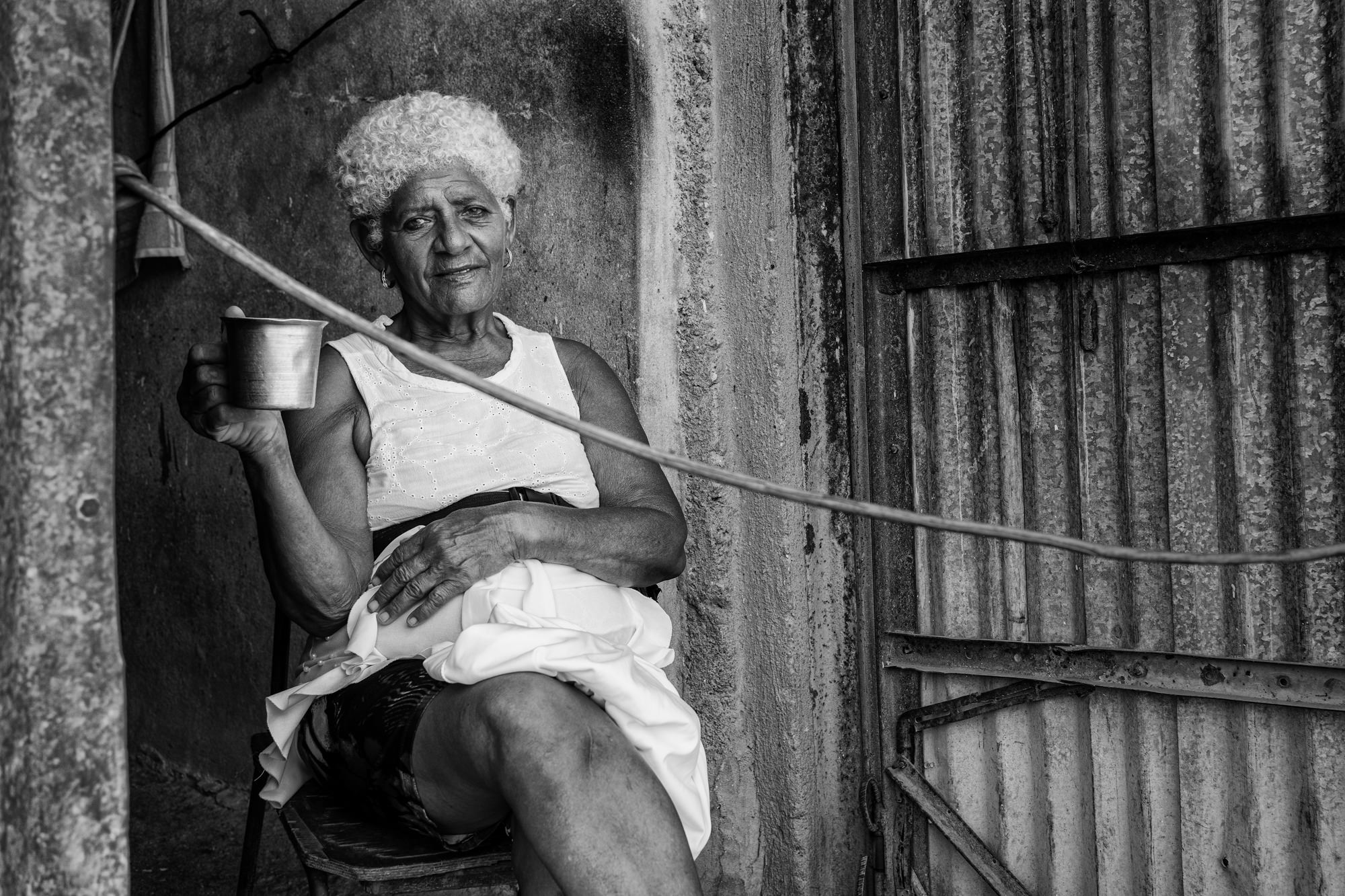 Cuba Street Photography Roadtrp | 14 Day Guide with Mark Seymour 28 “Elderly Cuban woman sitting in a doorway holding a metal cup in a quiet black-and-white moment.”