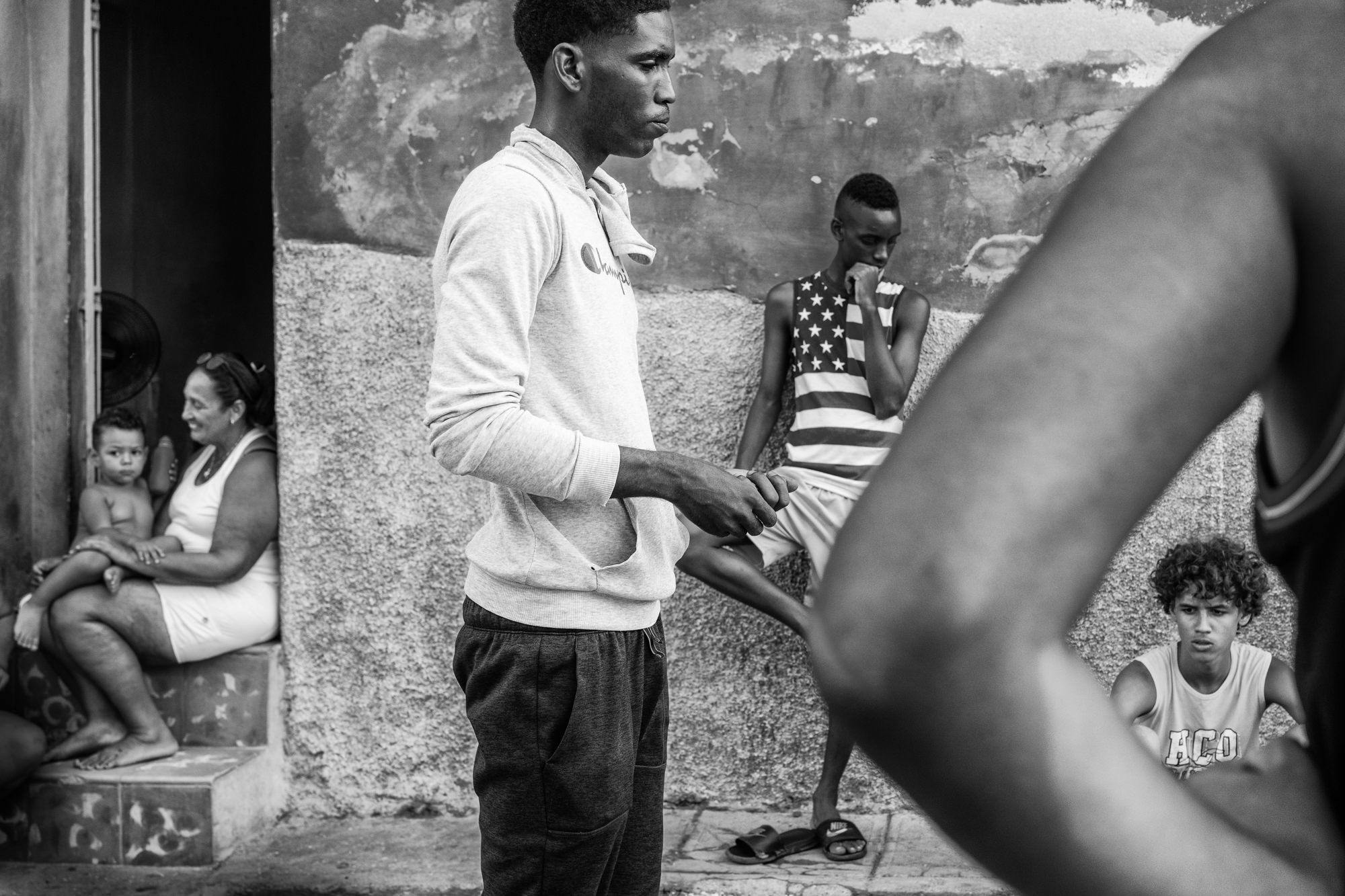 Cuba Street Photography Roadtrp | 14 Day Guide with Mark Seymour 25 “Black-and-white scene showing a young man in a hoodie standing near a wall as children and neighbours sit and watch from doorsteps.”