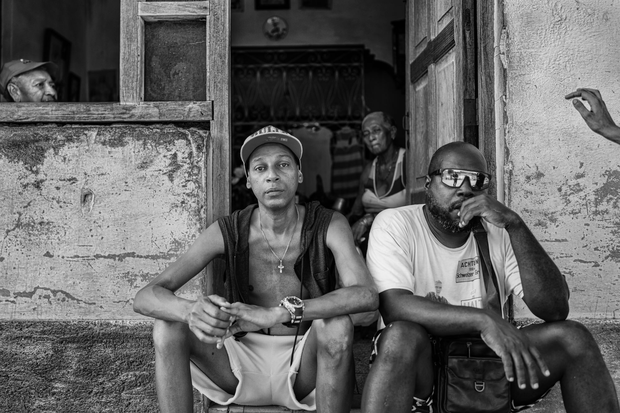 Cuba Street Photography Roadtrp | 14 Day Guide with Mark Seymour 23 “Two Cuban men sitting in a doorway in a black-and-white scene, with neighbours watching from inside the home.”