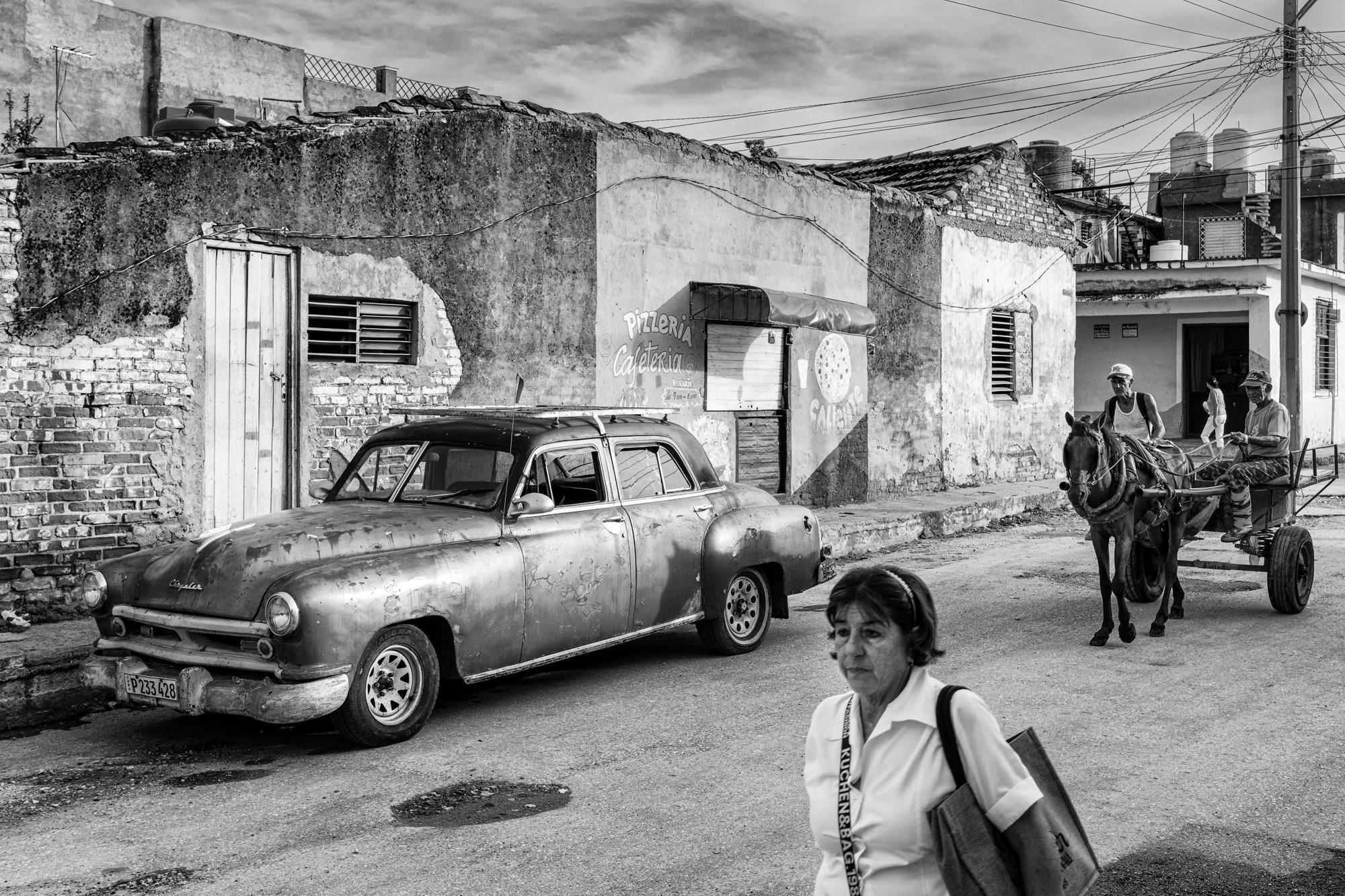 “Black-and-white Cuban street scene with a vintage car parked by a crumbling wall as a horse-drawn cart approaches.”