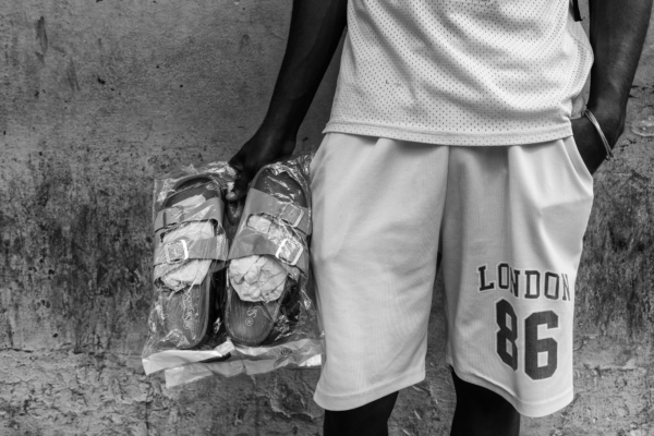 A close-up of a man in white shorts holding a pair of new sandals wrapped in plastic against a textured Cuban wall.