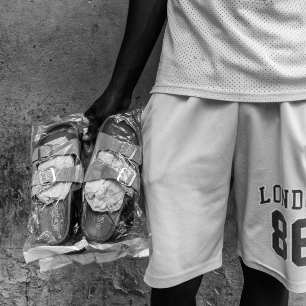 A close-up of a man in white shorts holding a pair of new sandals wrapped in plastic against a textured Cuban wall.