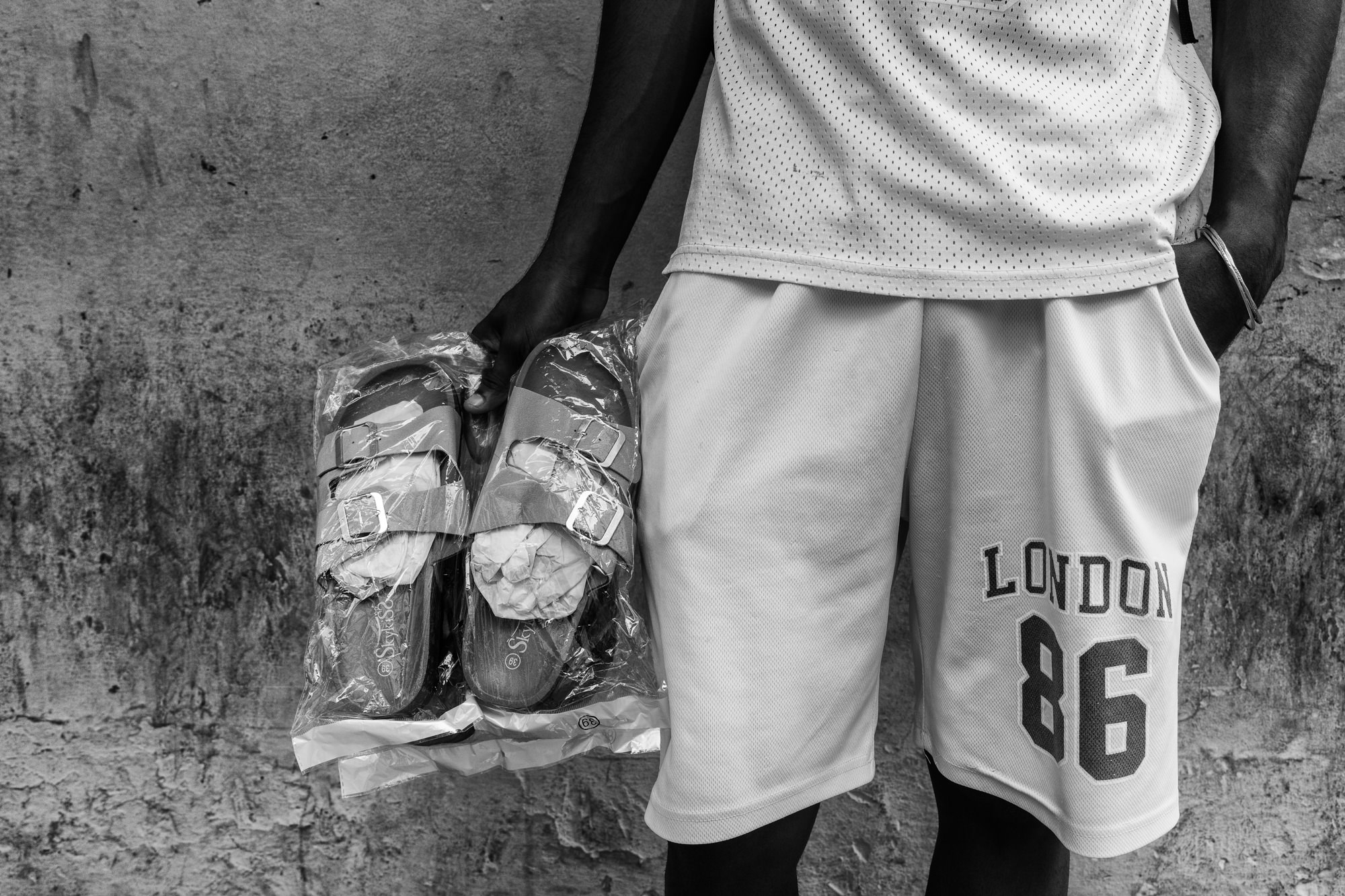 Cuba Street Photography Roadtrp | 14 Day Guide with Mark Seymour 15 A close-up of a man in white shorts holding a pair of new sandals wrapped in plastic against a textured Cuban wall.