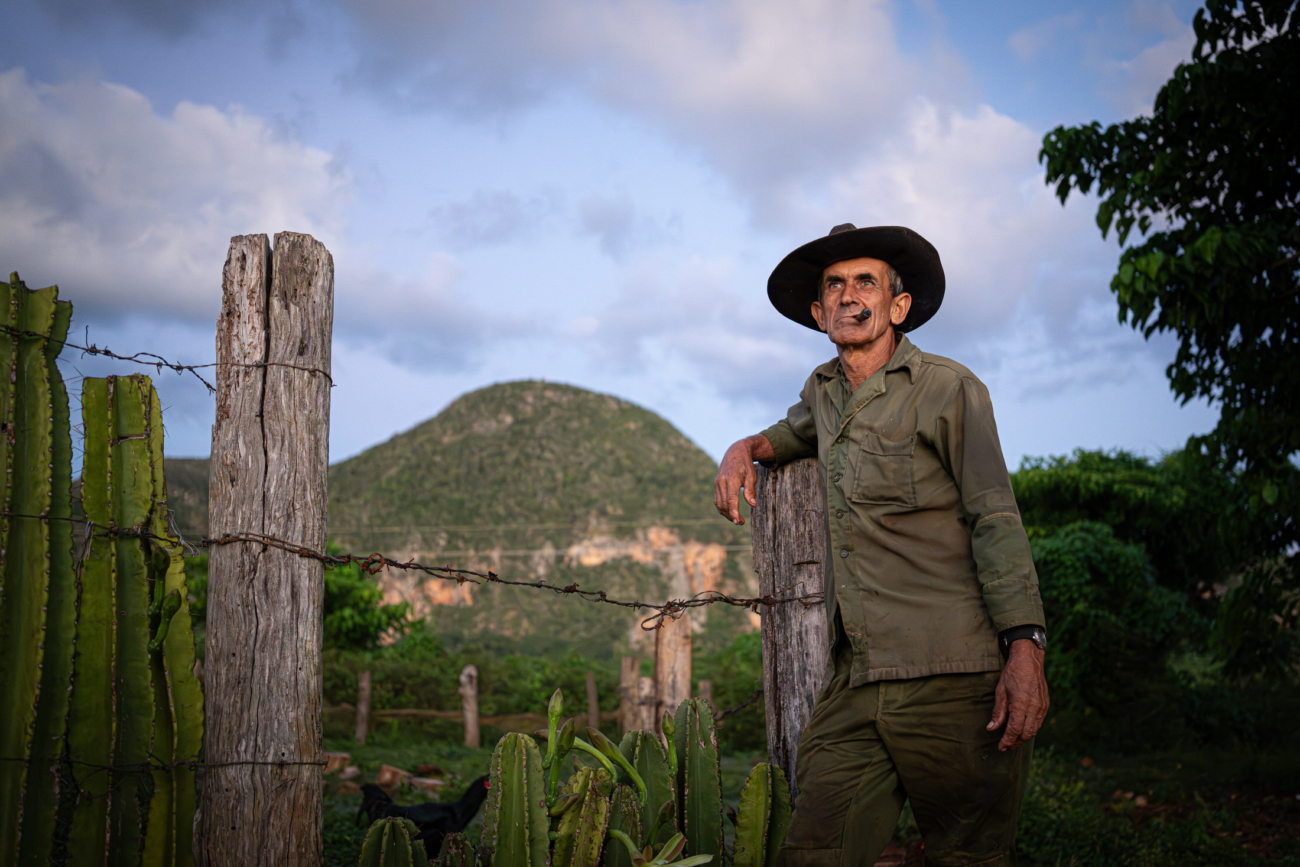 Cuba Street Photography Roadtrp | 14 Day Guide with Mark Seymour 6 A Cuban farmer in a wide-brimmed hat leaning on a wooden fence with lush green hills rising behind him at sunset.