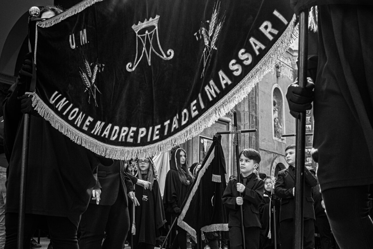 Inside the Misteri di Trapani: Documenting Sicily’s Living Tradition 14 A young boy stands beneath the banner of the Unione Madre Pietà dei Massari as musicians and participants wait outside the Church of the Anime Sante during the Misteri di Trapani in Sicily.