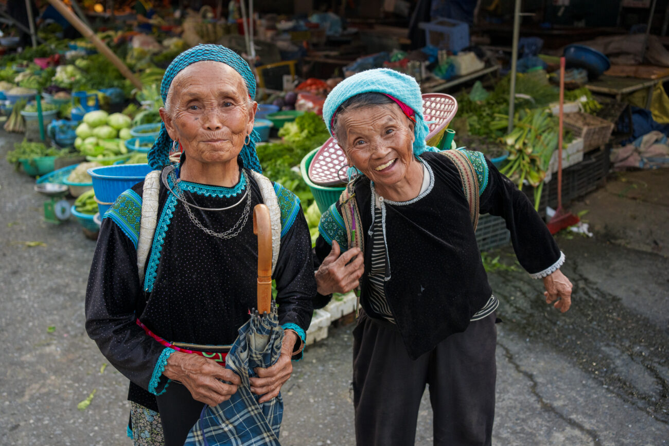Northern Vietnam Photography: A Photographer’s Journey Beyond the Tourist Trail 7 Two elderly women in traditional ethnic minority dress smile at the camera in a bustling market in northern Vietnam, surrounded by fresh vegetables and produce stalls.