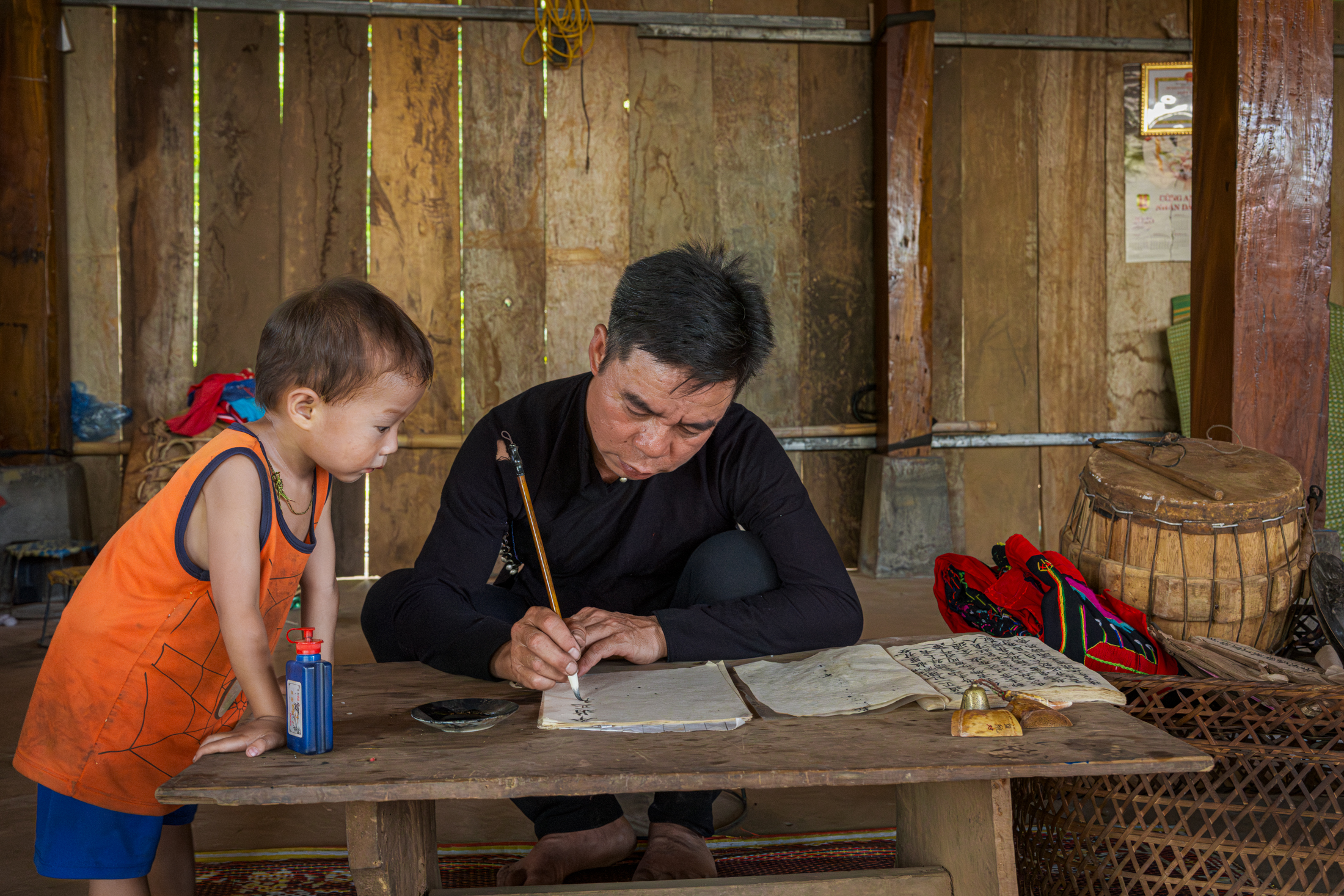 Northern Vietnam Photography: A Photographer’s Journey Beyond the Tourist Trail 17 A man writes traditional characters with brush and ink at a low wooden table while a young child watches closely inside a wooden house in northern Vietnam.