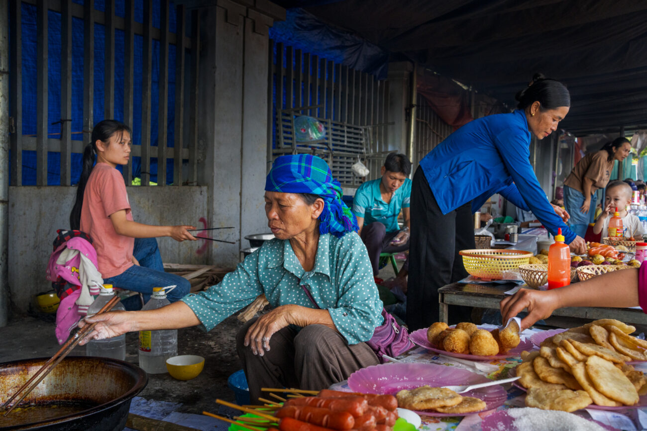 Northern Vietnam Photography: A Photographer’s Journey Beyond the Tourist Trail 8 A woman prepares fried snacks at a busy street food stall in a traditional market in northern Vietnam while vendors and customers gather around her.